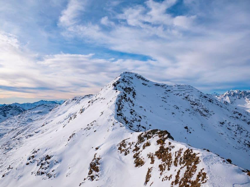 Una cima montuosa innevata e una cresta rocciosa sotto un cielo azzurro con nuvole bianche sparse.