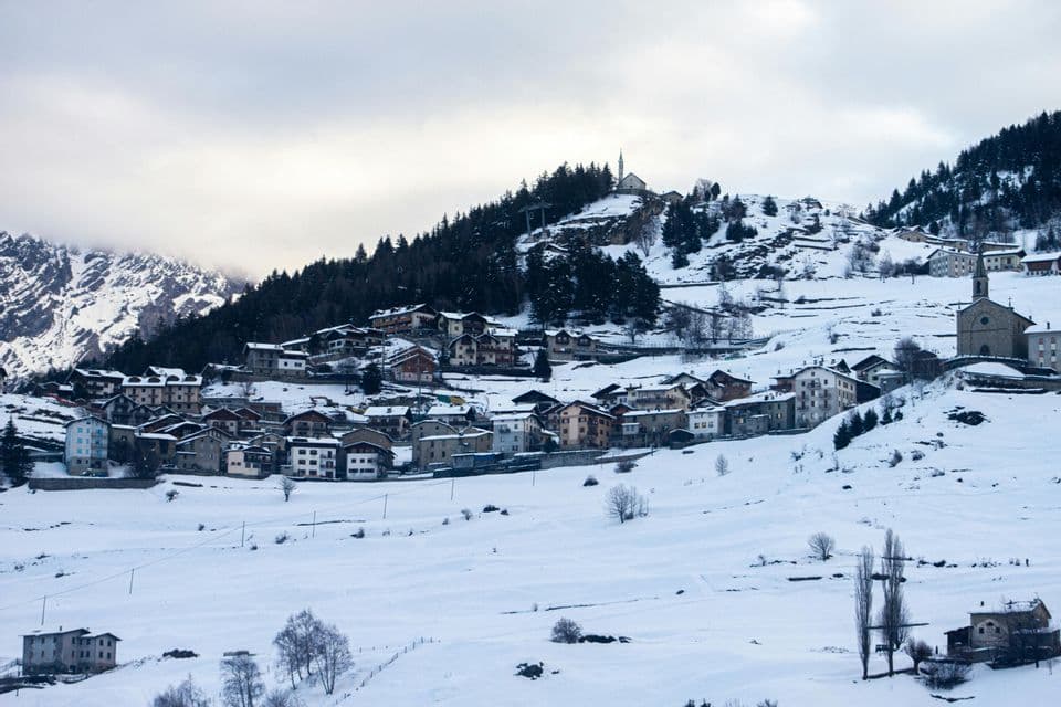 Un'ampia inquadratura di un villaggio costruito su un fianco di montagna innevato, punteggiato da pini, sotto un cielo nuvoloso.