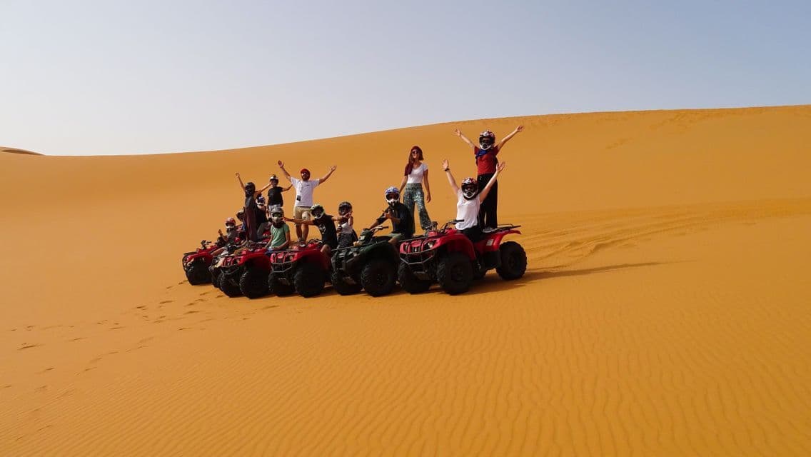 A WeRoad group trip posing on red and green quad bikes, some with arms raised, in a vast desert with orange sand dunes.