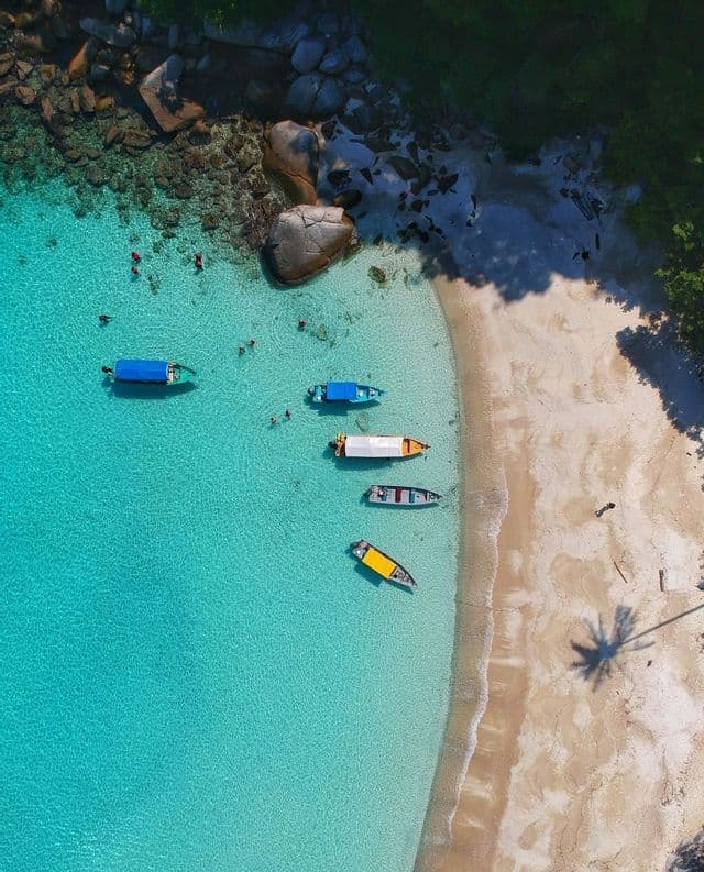 Una vista aérea de una cala tropical con gente nadando y barcos amarrados en aguas cristalinas de color turquesa junto a una playa de arena.