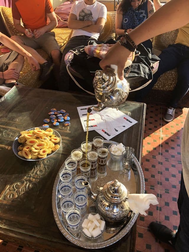 A person pours tea from an ornate silver teapot into small glasses for a WeRoad group trip seated around a table.