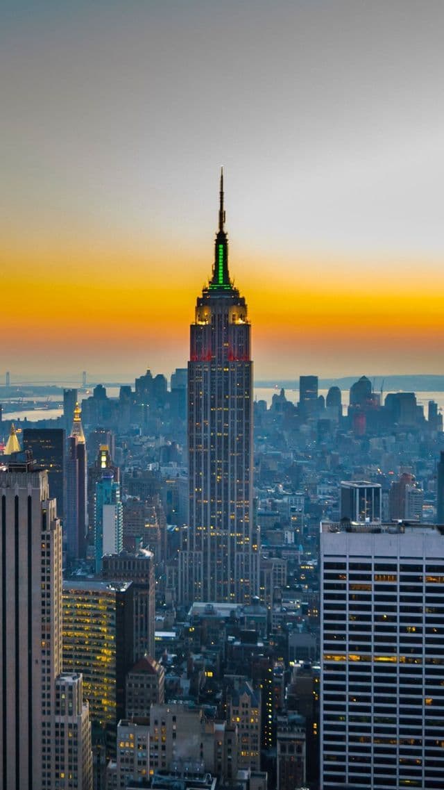 Das Empire State Building ragt bei Sonnenuntergang über einer dichten Stadtsilhouette empor, seine Turmspitze leuchtet grün und rot vor einem orangefarbenen Himmel.