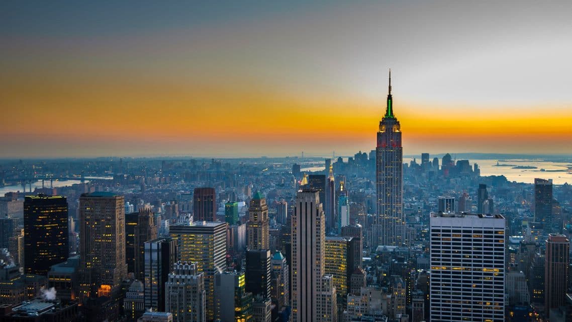 An expansive city skyline with numerous illuminated skyscrapers, including a prominent tower, against a vibrant sunset.