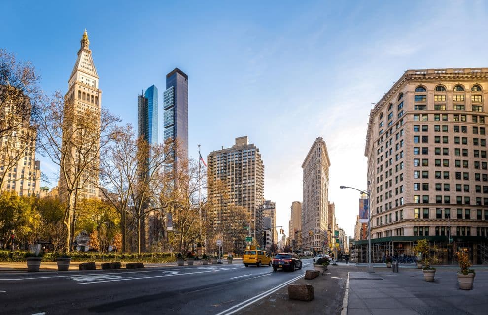 A wide city street view with the Flatiron Building and other skyscrapers under a clear, sunny sky with light traffic.