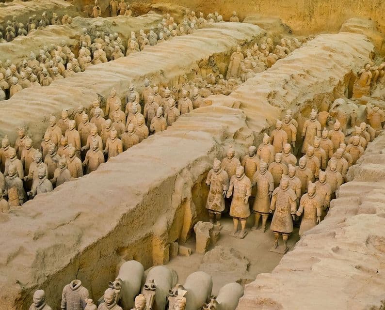 Rows of life-sized terracotta warrior statues standing in formation inside large, excavated earthen pits.