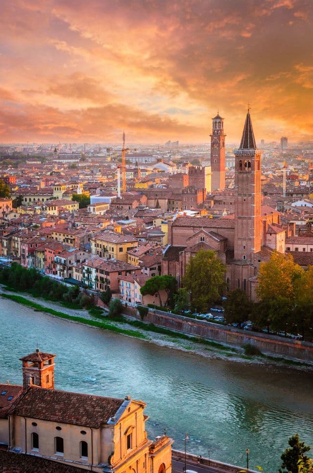 Una veduta aerea di una città storica con tetti in terracotta e alte torri campanarie lungo un fiume sotto un tramonto dorato.