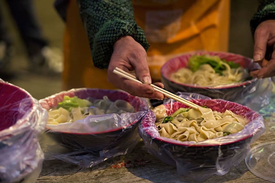 A person uses chopsticks to serve flat noodles and greens into bowls lined with plastic at a street food stall.