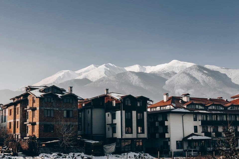 Alpine-style buildings with snowy roofs stand before a vast, snow-capped mountain range under a clear sky.