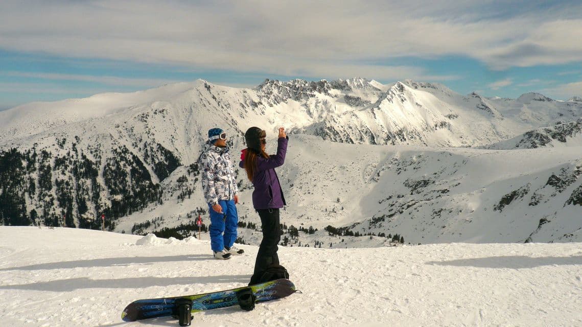 Dos snowboarders están de pie en una pendiente nevada, y uno de ellos toma una foto de la vasta cordillera frente a ellos.