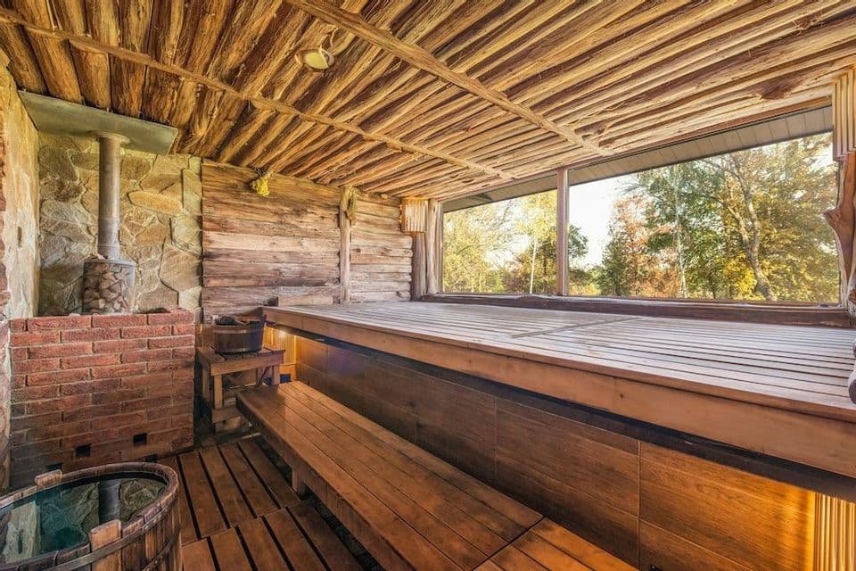 El interior de una sauna de madera rústica con una estufa de piedra, bancos en niveles y una gran ventana con vistas al bosque.