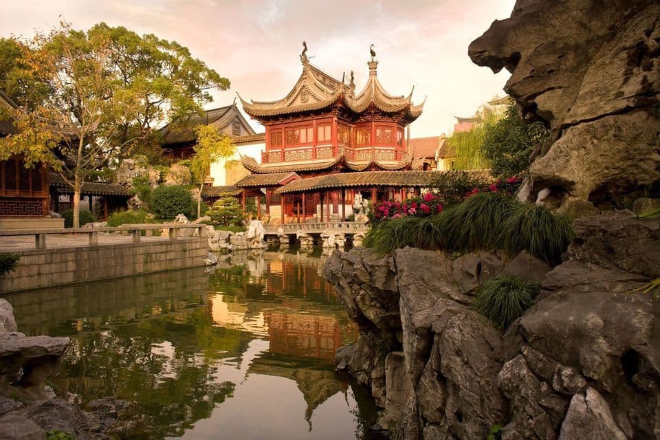 A traditional red Chinese pavilion with tiered roofs reflects in a calm pond, surrounded by lush gardens and rocks.