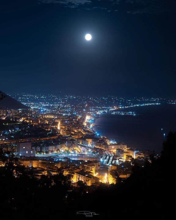 Una luna llena cuelga en el cielo oscuro sobre una extensa ciudad costera iluminada por luces amarillas y azules por la noche, vista desde un mirador elevado.