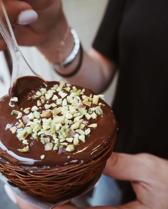 Primo piano di un dolce a strati, ricoperto di glassa al cioccolato e pistacchi tritati, che viene mangiato con un cucchiaio di plastica trasparente.