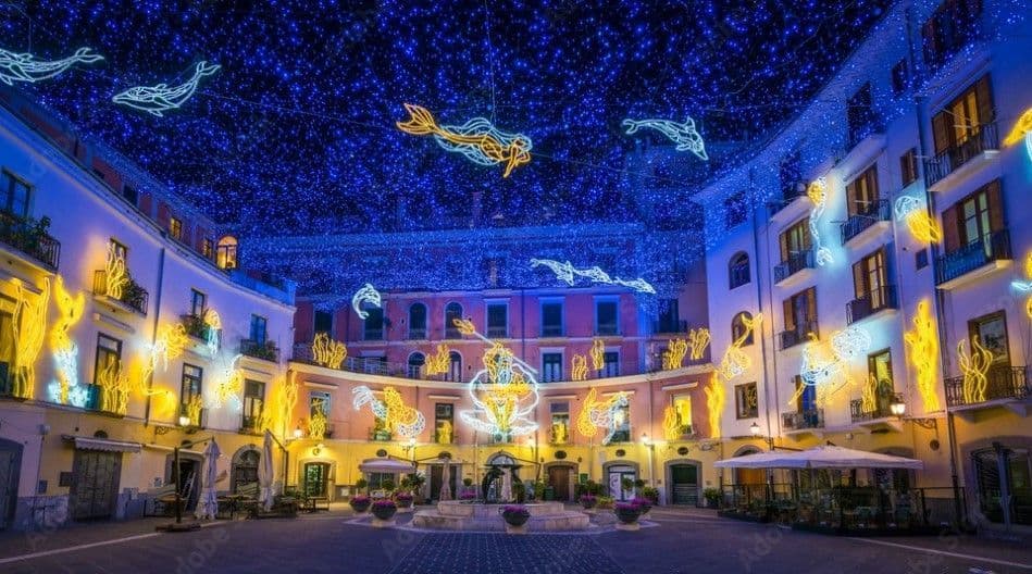Una plaza de noche decorada con instalaciones lumínicas de temática marina, incluyendo sirenas y peces, bajo un dosel de luces azules.