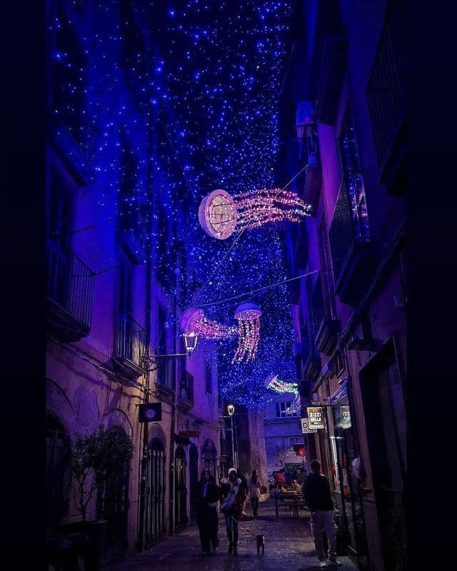 Lanterne illuminate a forma di medusa e lucine blu pendono su una stretta strada acciottolata di notte.