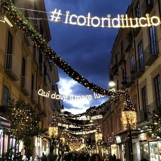 Letreros luminosos y guirnaldas festivas cuelgan sobre una calle estrecha y concurrida al anochecer, con edificios a ambos lados.