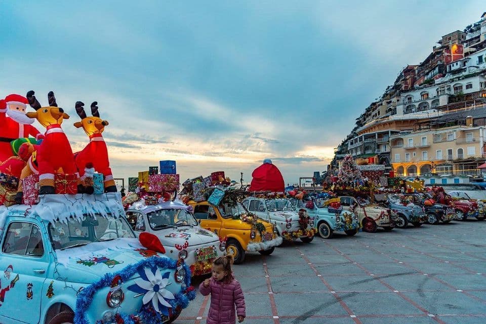 Una fila de Fiat 500 clásicos, adornados con elaboradas decoraciones navideñas, estacionados en una plaza al anochecer.
