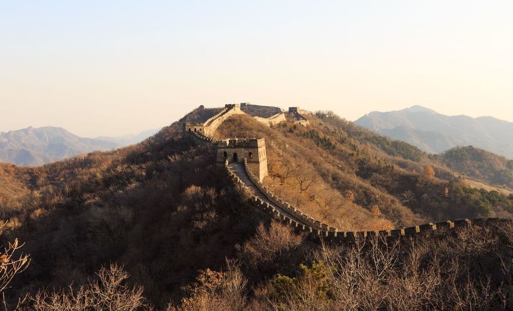 La Gran Muralla China serpentea por colinas ondulantes y cubiertas de árboles, con una prominente atalaya bajo un cielo pálido y brumoso.