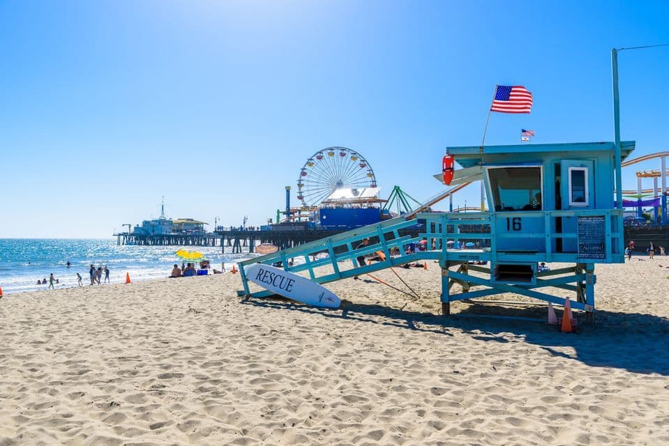 A blue lifeguard tower on a sunny beach, with a pier and a Ferris wheel in the background under a clear sky.