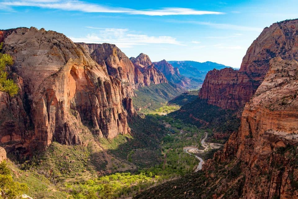 A high-angle view of a vast red rock canyon with a lush green valley and a winding river below, under a blue sky.