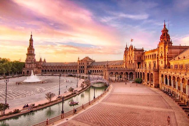 Un edificio decorato con torri e arcate circonda un'ampia piazza con un canale e una fontana sotto un cielo al tramonto colorato.