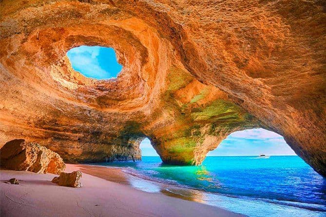 Una vista dall'interno di una grotta marina di roccia dorata con spiaggia sabbiosa, che si apre sull'oceano turchese e il cielo blu attraverso un oculo.