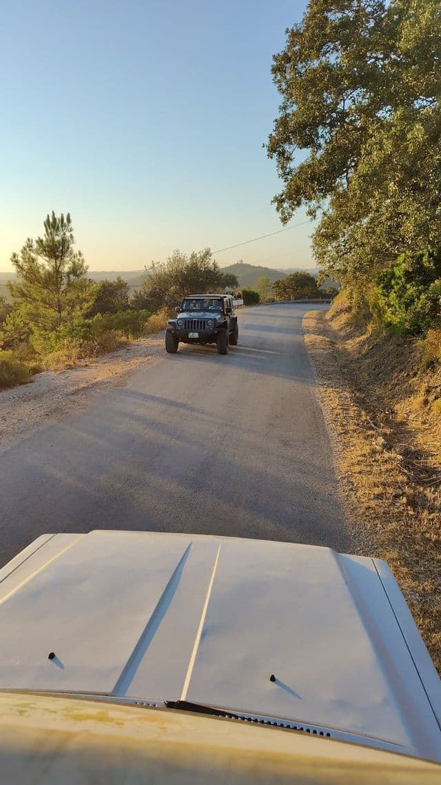 Un viaggio di gruppo WeRoad in Jeep nera su una strada tortuosa attraverso un paesaggio collinare illuminato dal sole, visto dal cofano di un altro veicolo.