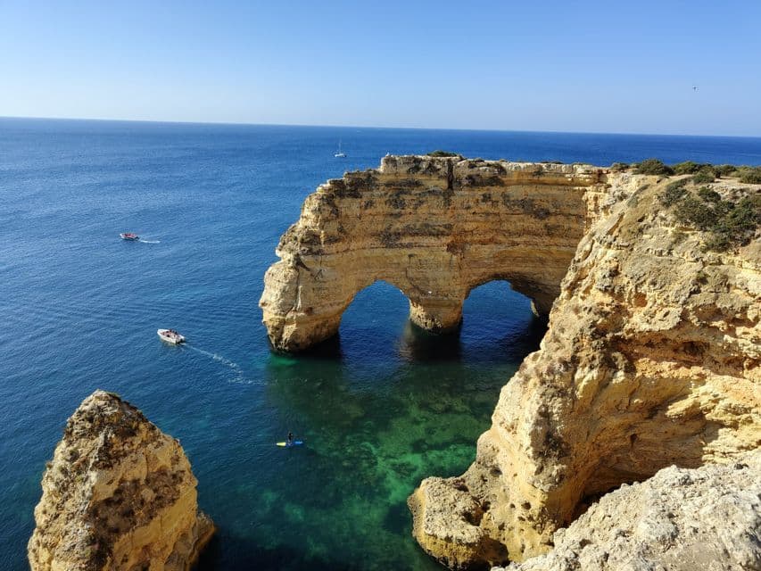 Una costa rocciosa presenta una doppia formazione ad arco marino in acque limpide e blu, con piccole imbarcazioni e una persona su un paddleboard nelle vicinanze.