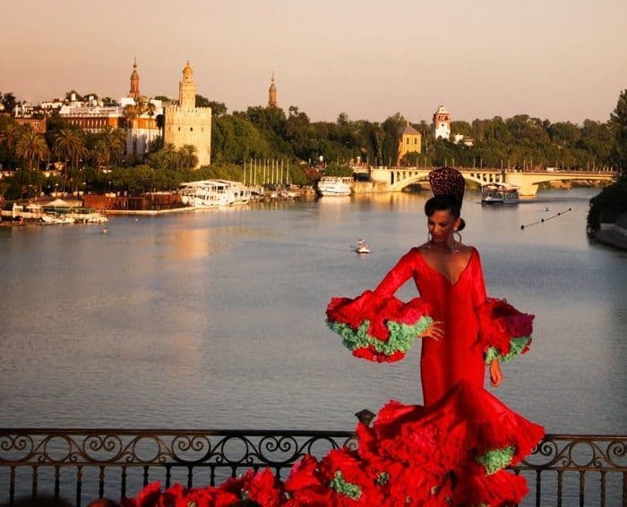 Una donna in abito da flamenco rosso posa su un balcone che si affaccia su un fiume e una città storica al tramonto.