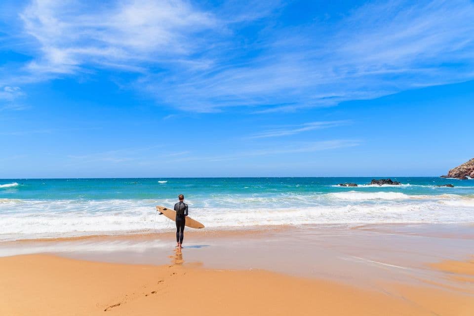 Un surfista in muta con una tavola da surf sta su una spiaggia sabbiosa guardando l'oceano turchese sotto un cielo blu.