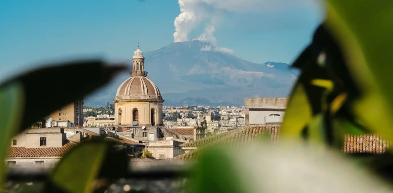 An erupting volcano with a smoke plume rises above a cityscape with a church dome, framed by green leaves.