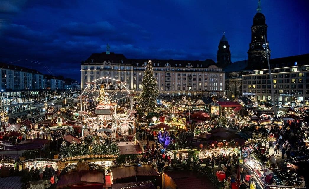 An aerial view of a bustling Christmas market in a city square at night, with an illuminated Ferris wheel and a large Christmas tree.