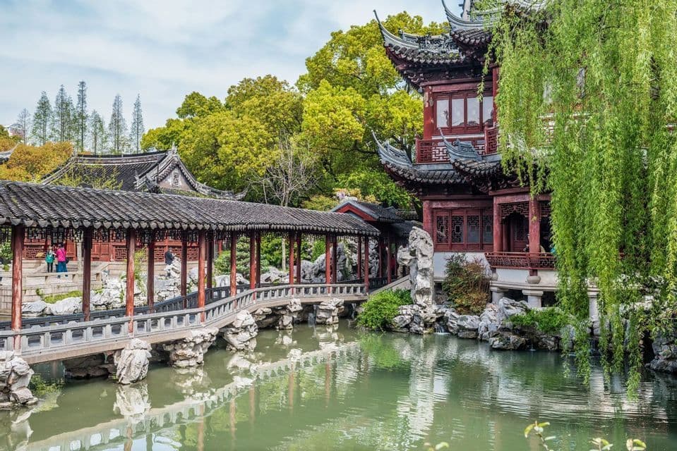 A traditional Chinese pavilion and covered zigzag bridge cross a tranquil pond in a lush classical garden with a weeping willow.