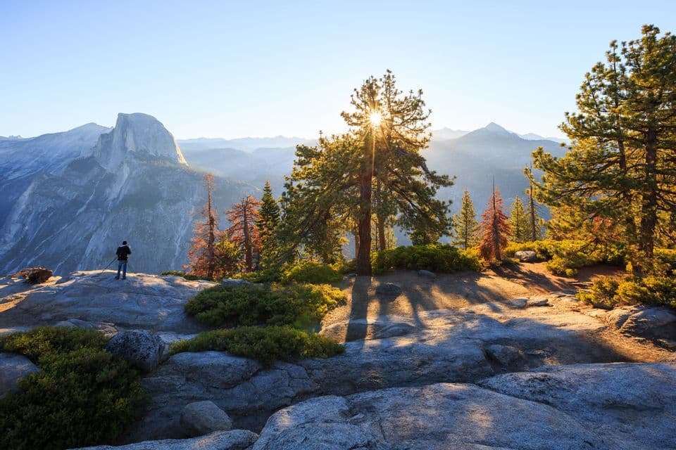 Sonnenlicht strömt durch Kiefern, während ein Fotograf auf einem felsigen Aussichtspunkt den Blick auf einen Granitberg einfängt.