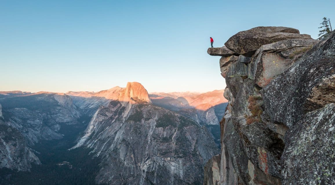 Une personne en veste rouge se tient sur une étroite corniche rocheuse, surplombant une vaste chaîne de montagnes éclairée par le soleil matinal.