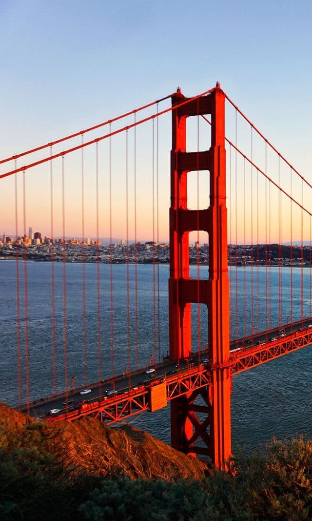 The red-orange tower of the Golden Gate Bridge at sunset, with traffic on the deck and a city skyline in the background across the water.