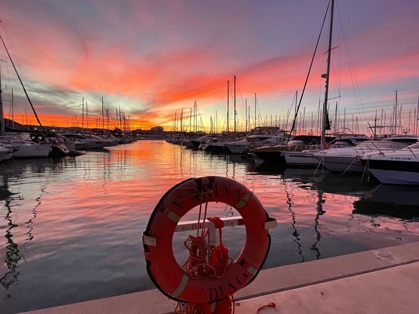 Un vibrante atardecer naranja y morado sobre una marina llena de veleros, con un salvavidas rojo en el muelle en primer plano.