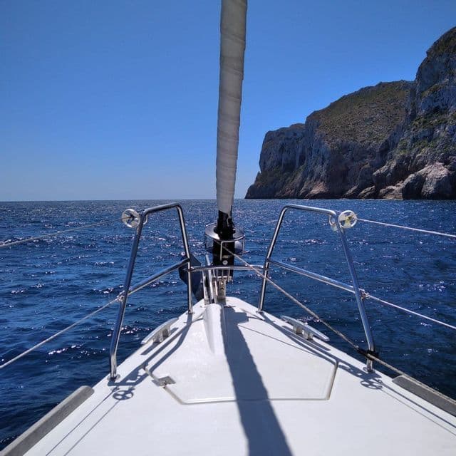 Vista desde la proa de un velero navegando por el mar azul hacia un acantilado rocoso.