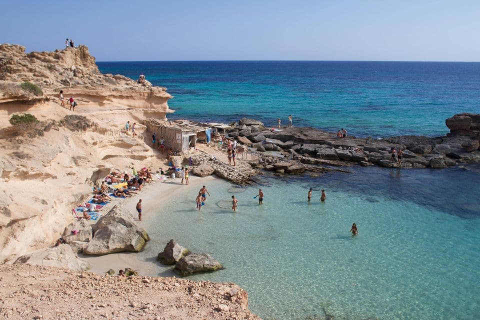 Vista elevada de una cala rocosa y soleada donde la gente toma el sol en una pequeña playa y nada en agua clara y turquesa.