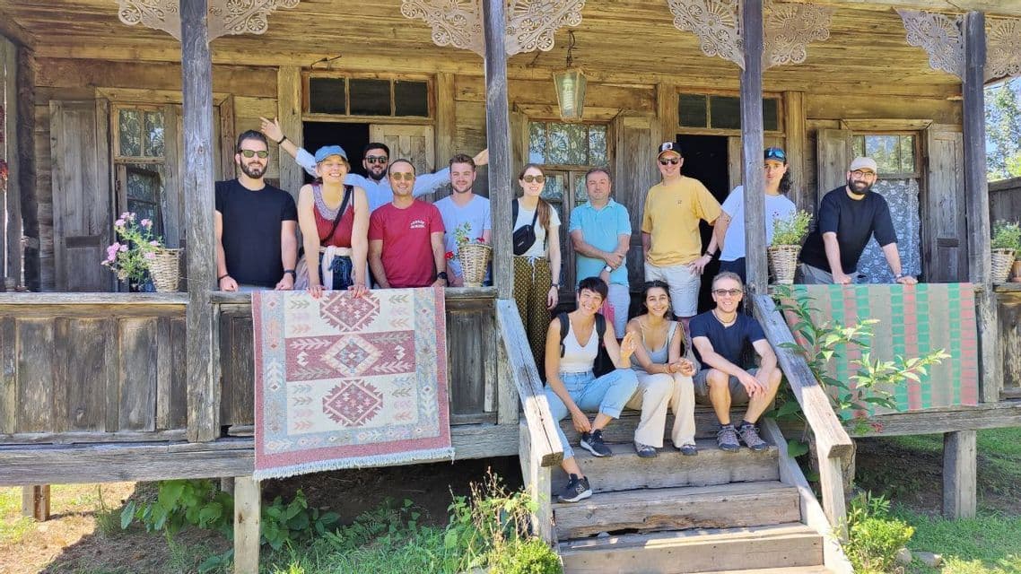 Un gruppo WeRoad in posa per una foto sul portico di una casa rustica in legno, mentre tiene in mano un tappeto a fantasia.