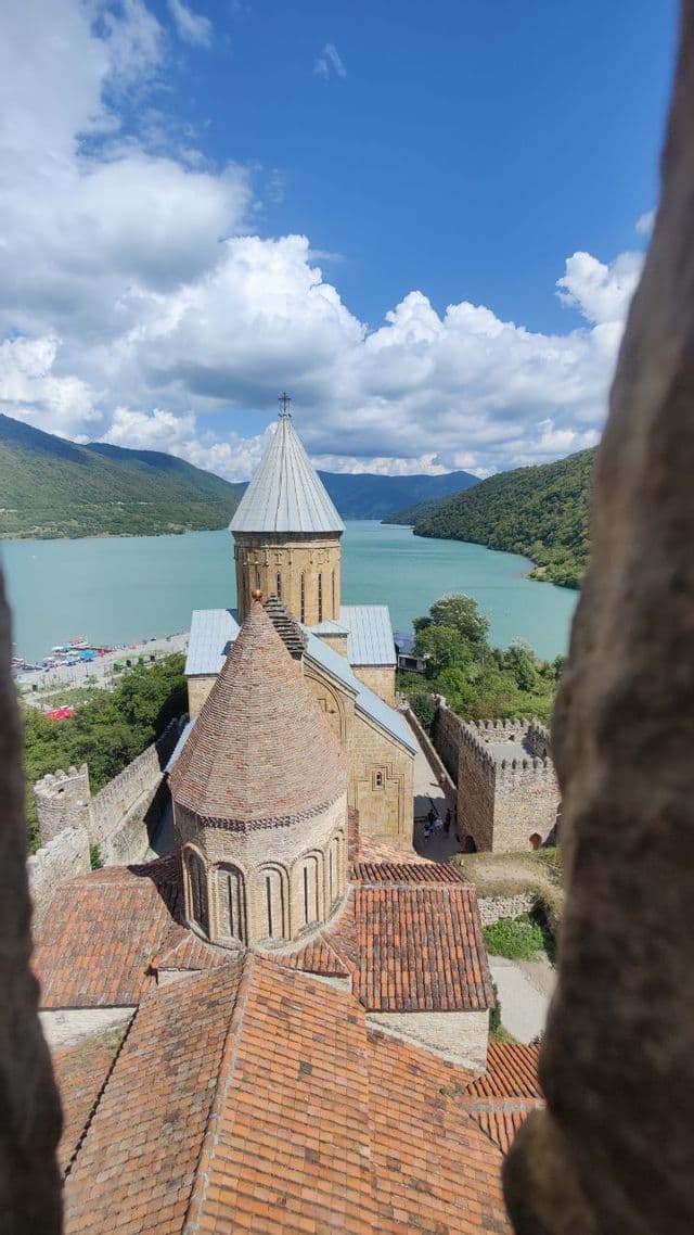 Una fortezza di pietra con tetti conici, vista da un'altura, domina un lago turchese e colline boscose sotto un cielo nuvoloso.