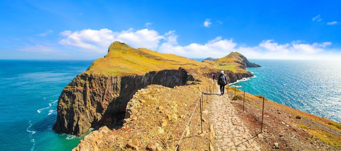 A person with a backpack hikes along a narrow stone path on a cliff's edge, overlooking a vast blue ocean under a clear sky.