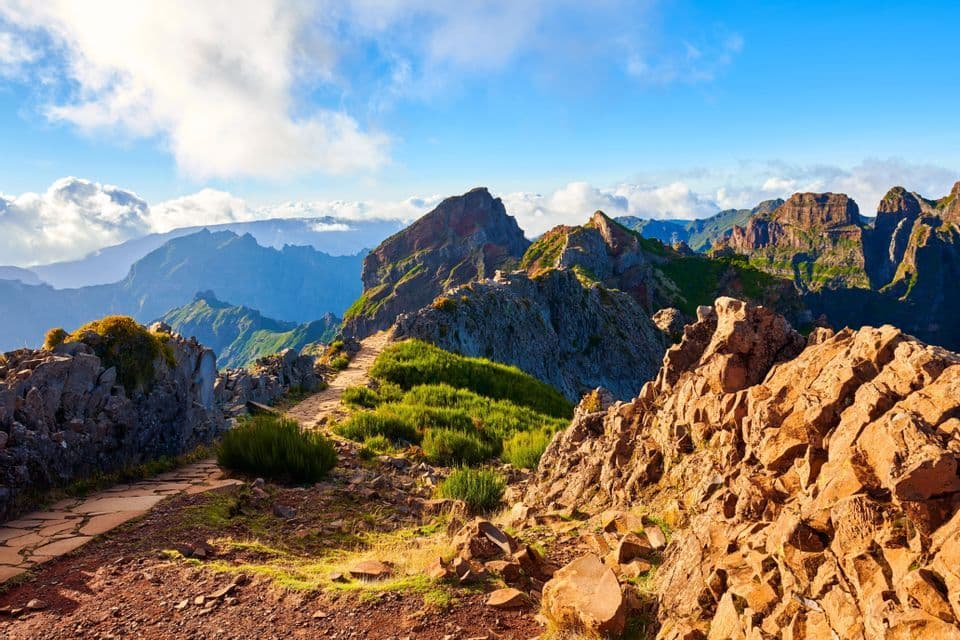 A stone path winds along a rugged mountain ridge with expansive views of peaks and clouds under a blue sky.