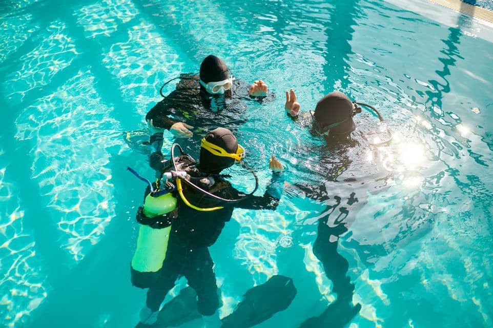 Trois participants d'un voyage en groupe WeRoad, équipés de matériel de plongée, s'exercent aux signaux manuels pendant un cours en piscine.