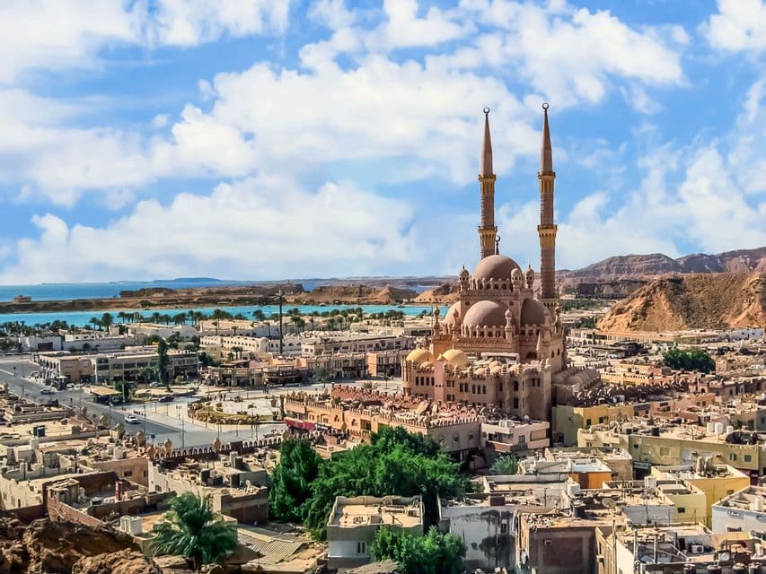 Une grande mosquée avec deux minarets et de multiples dômes surplombe une ville côtière aux eaux turquoise et aux collines arides à l'horizon.