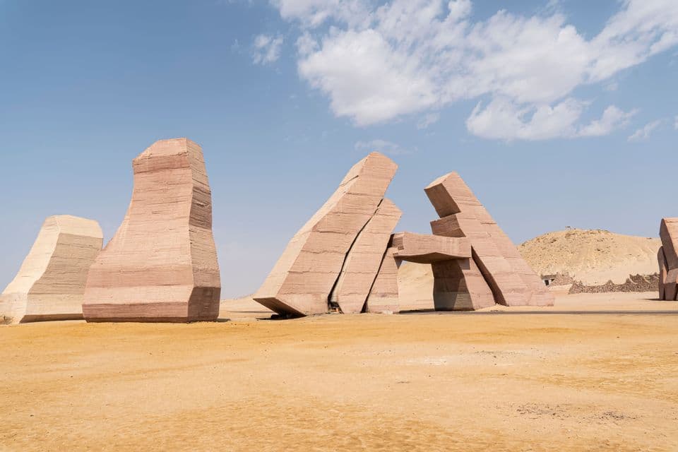 De grandes sculptures angulaires en pierre stratifiée et colorée se dressent sur le sable d'un désert sous un ciel partiellement nuageux.