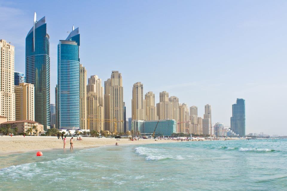 Una spiaggia di sabbia con acqua turchese incontra un denso skyline urbano di moderni grattacieli sotto un cielo limpido e luminoso.