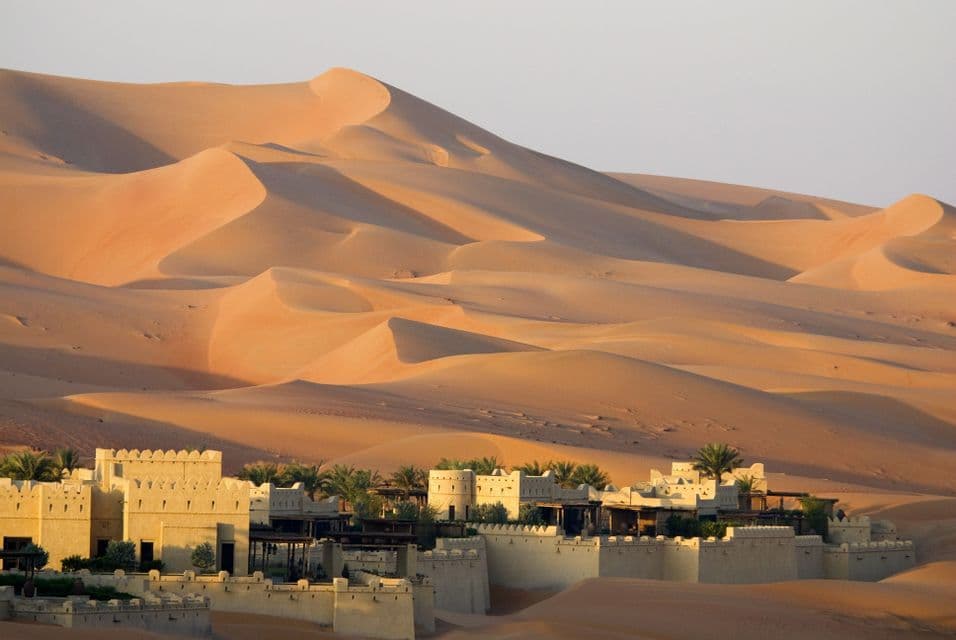 Un complejo fortificado con palmeras se encuentra entre vastas dunas de arena naranja al amanecer.