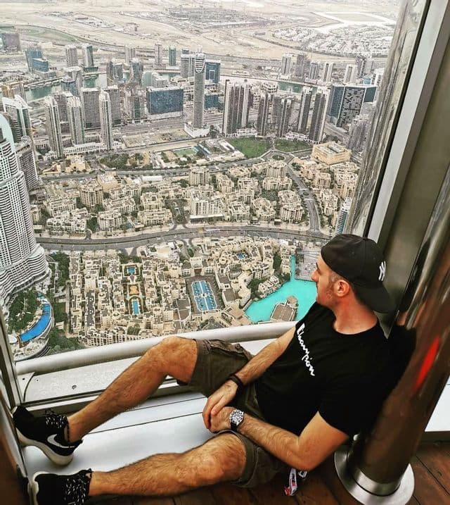 Un hombre con gorra se sienta en el suelo junto a una ventana, contemplando un extenso paisaje urbano desde un punto de vista elevado.