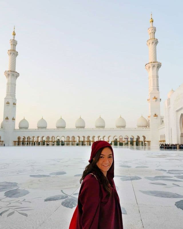 Una donna con veste rossa e cappuccio sorride nel grande cortile di una moschea bianca con due minareti.